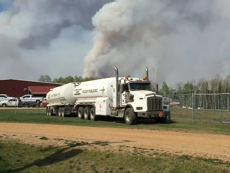 A Flight Fuels tanker truck parked at a base with a wildfire smoke column rising behind